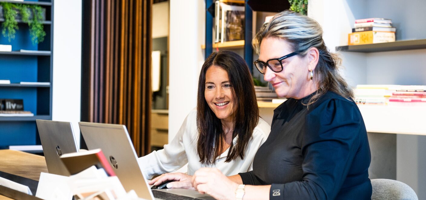 afbeelding van twee vrouwen ieder achter een laptop. een vrouw kijkt lachend bij de andere vrouw op de laptop