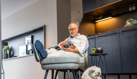 man zittend in een stoel en leest tijdschrift. naast hem op de grond ligt een klein hondje.