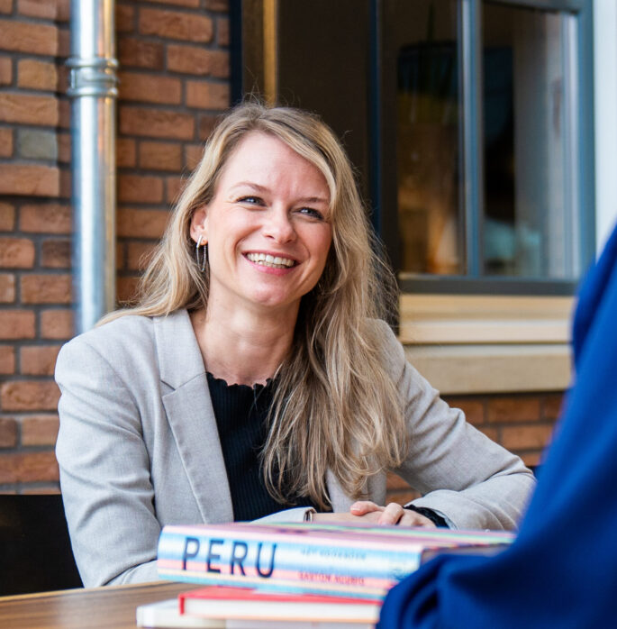 foto van medewerker die lachend aan een tafel zit met een stapel boeken voor zich